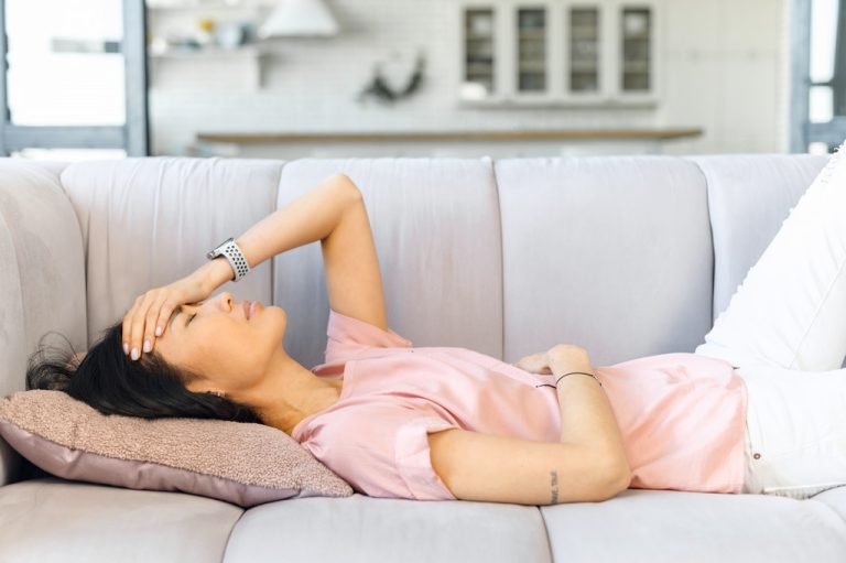 Young Asian Woman Laying On a Couch Holding Her Stomach and Head Experiencing Common Pregnancy Symptoms