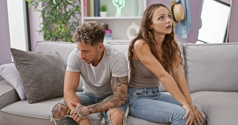Young couple sitting back-to-back on a couch, appearing thoughtful and uncertain about an unexpected pregnancy.
